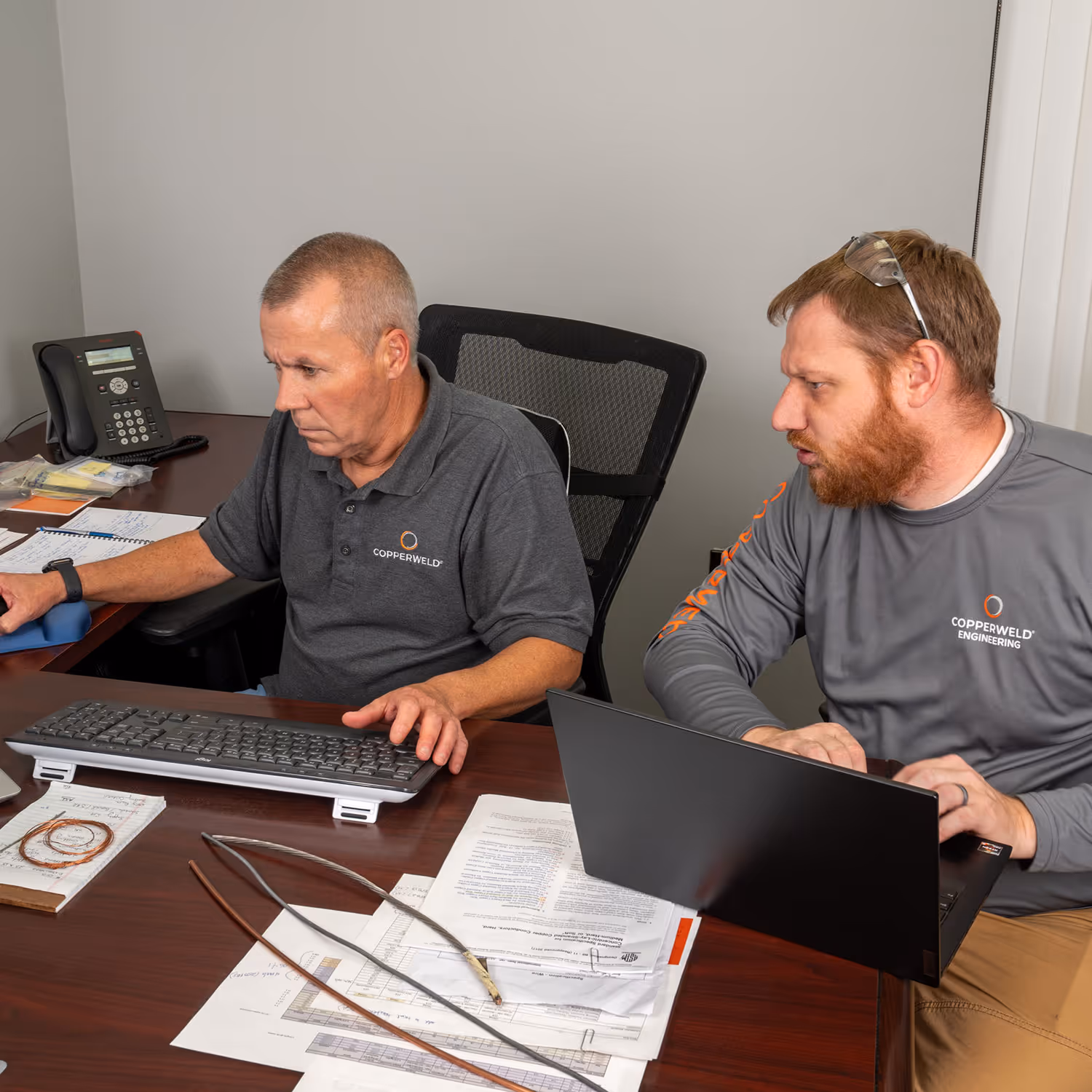 Two men in Copperweld shirts work at a desk with computer, documents, and metal wire samples.