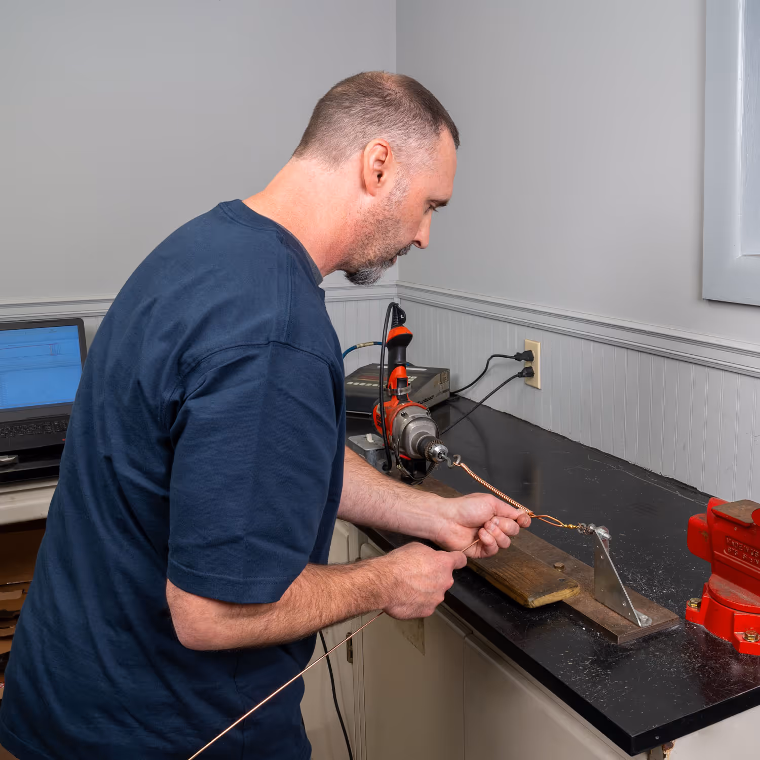 Man using a drill to twist copper wire held on a workbench setup with a red vise and laptop nearby.