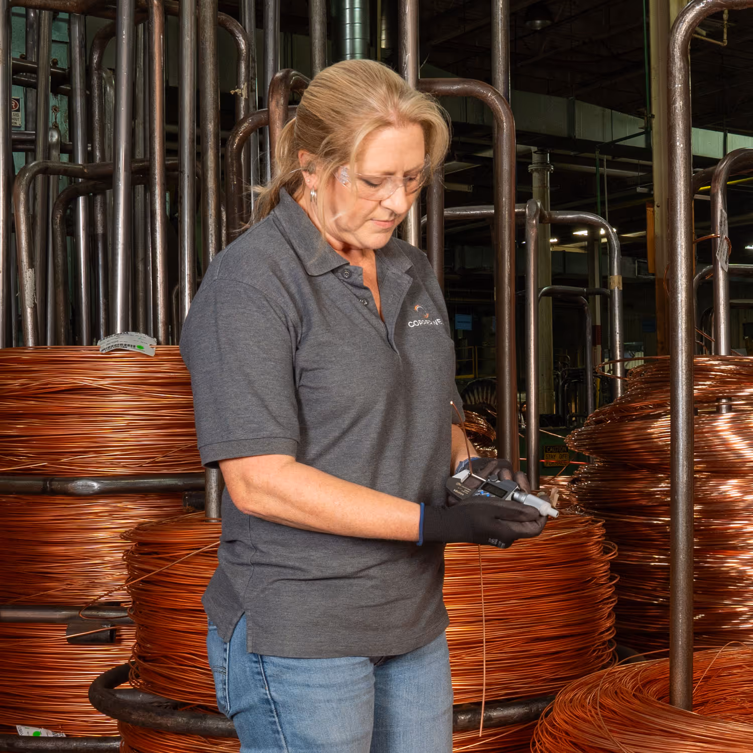 Woman in safety glasses and gloves inspecting copper wire coils in an industrial factory setting.