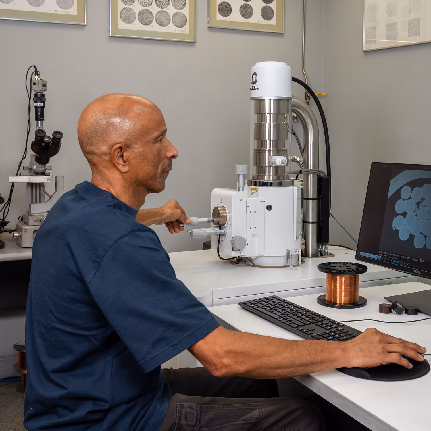 Man operating a scientific electron microscope with a spool of copper wire and monitor showing sample details.