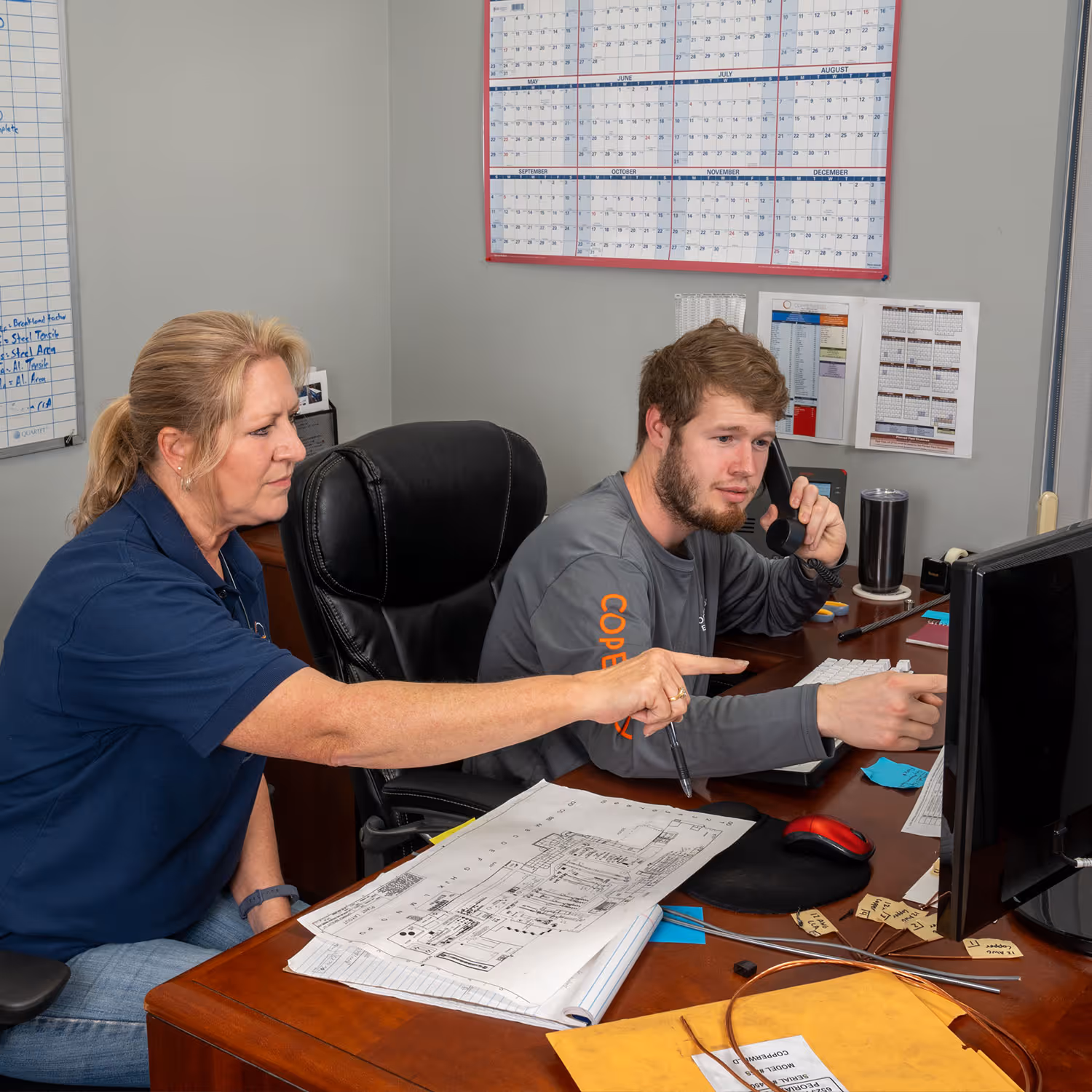 A man on the phone and a woman pointing at a computer screen in an office with documents and a calendar.