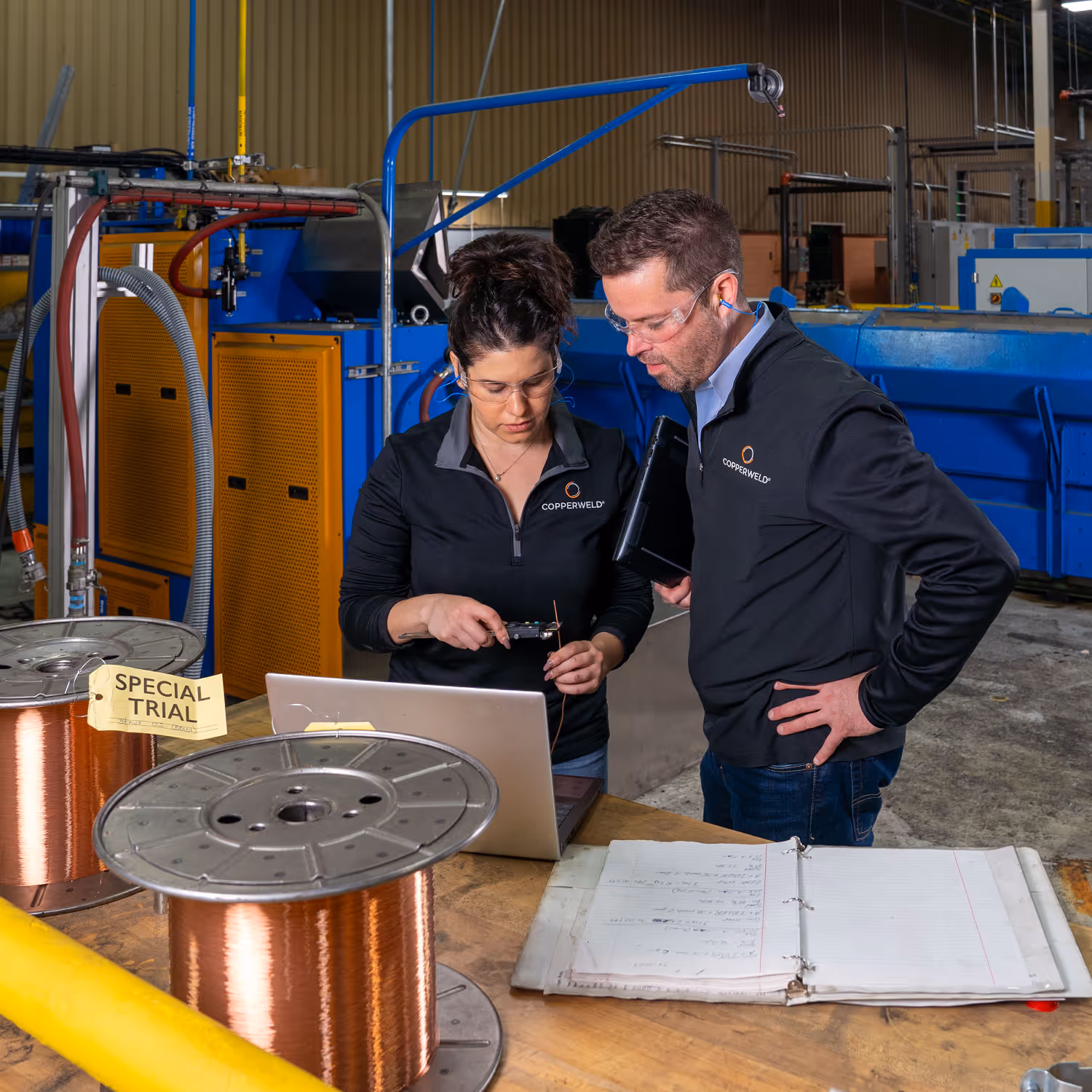 Two factory workers in safety glasses inspecting copper wire near laptop and notes in industrial setting.
