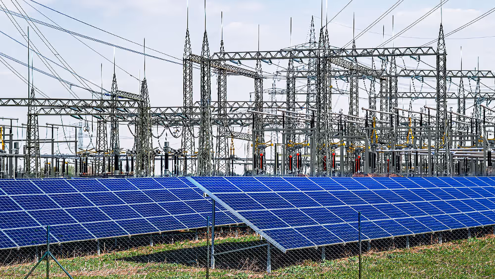 Rows of blue solar panels in front of an electrical substation with power lines and towers.