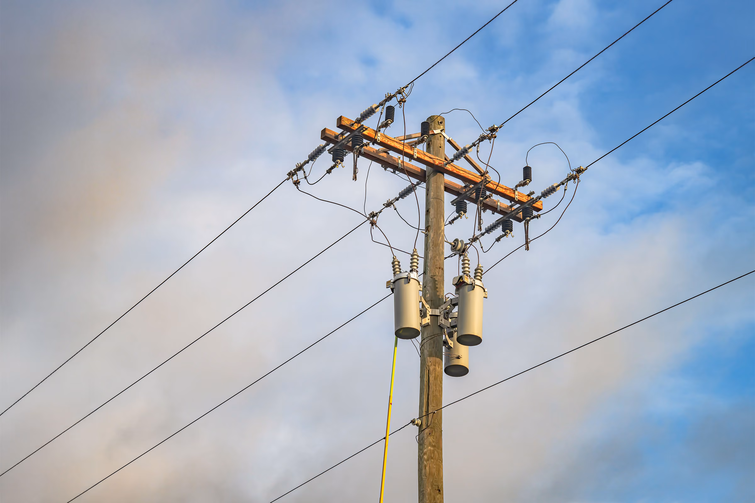 Wooden power pole with transformers and multiple electrical wires against a partly cloudy blue sky.