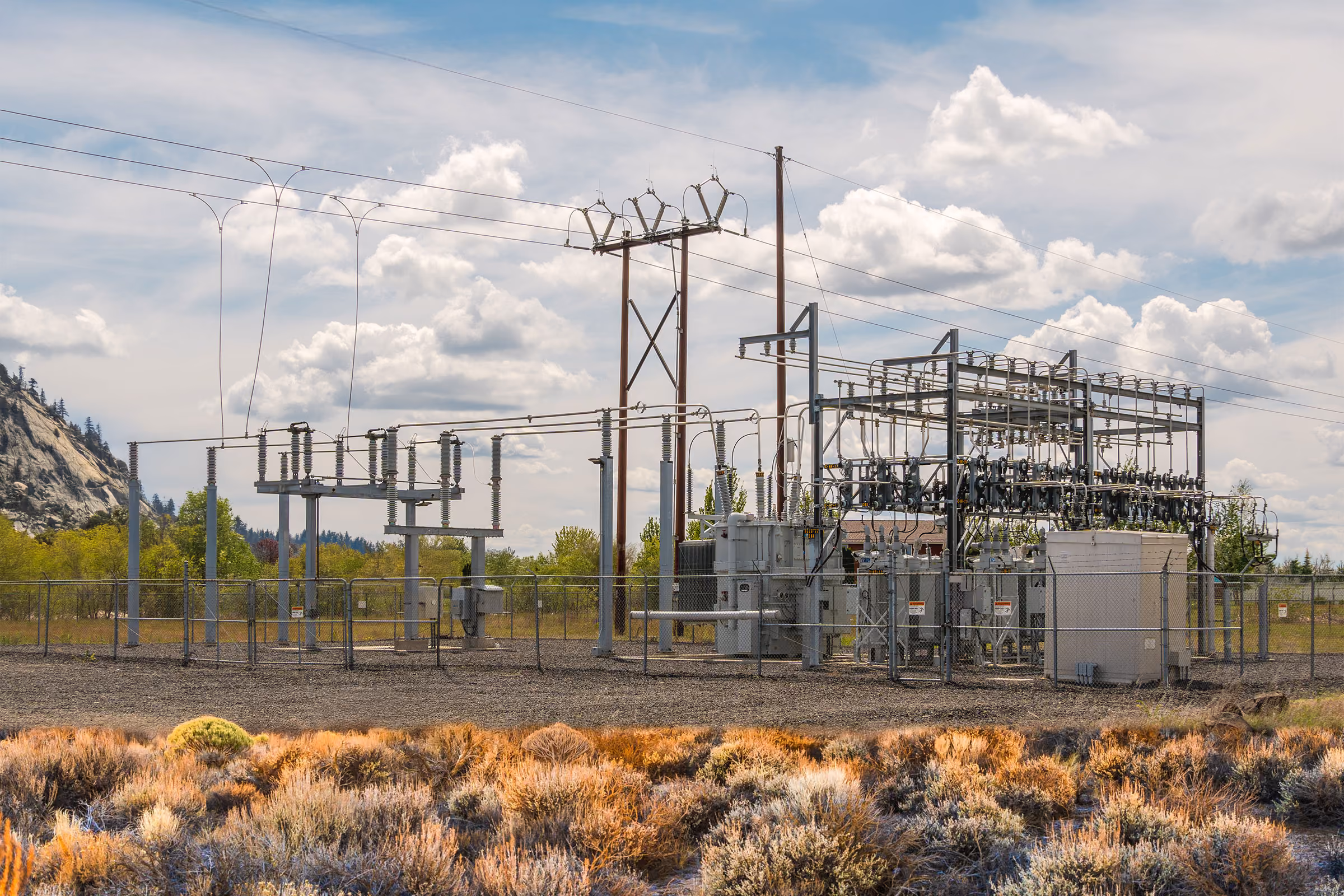 Electrical substation with transformers and power lines set against a partly cloudy sky and natural landscape.