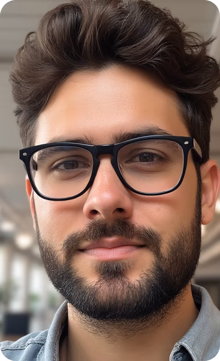 Close-up of a man with dark hair, beard, and black rectangular glasses, wearing a denim shirt indoors.