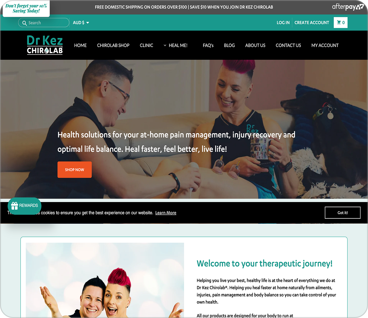 Two smiling women sitting on a couch with one applying therapy equipment on the other's leg while a dog rests beside them.