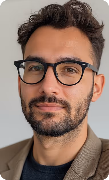 Close-up of a man with dark hair and beard wearing black framed glasses and a brown blazer against a plain background.