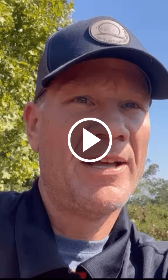 Man wearing a black cap and jacket speaking outdoors in front of green trees and blue sky.