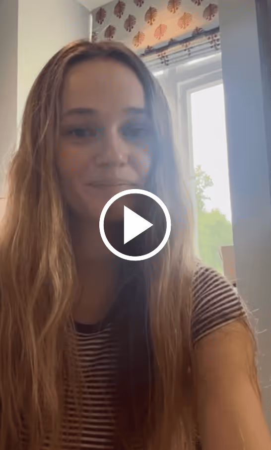 Young woman with long wavy hair and striped shirt sitting indoors near a window with light curtain and leafy wallpaper above.