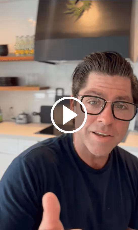 Man wearing black glasses and a dark shirt in a kitchen setting, gesturing with his hand toward the camera.