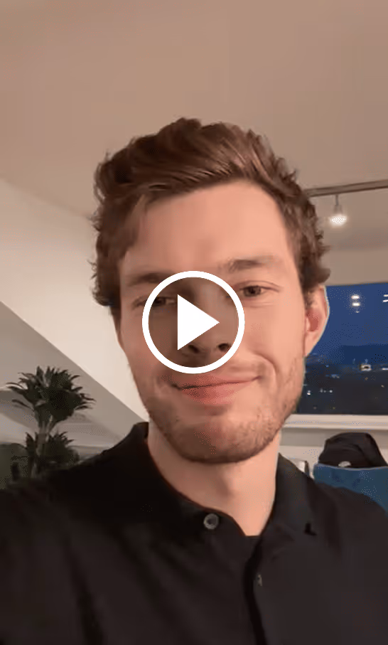 Young man with short curly hair and a beard smiling indoors with houseplants and a window in the background.