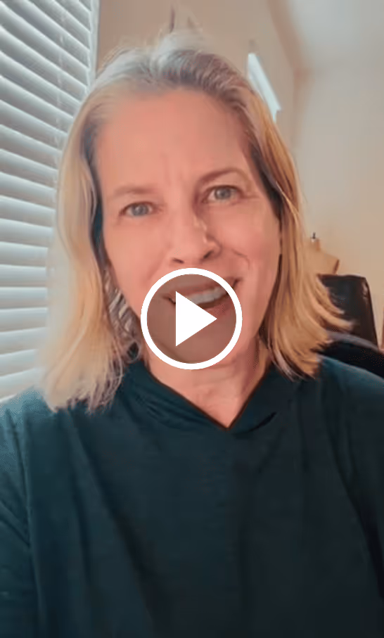 Smiling woman with shoulder-length blonde hair wearing a dark hoodie, sitting indoors near window blinds.