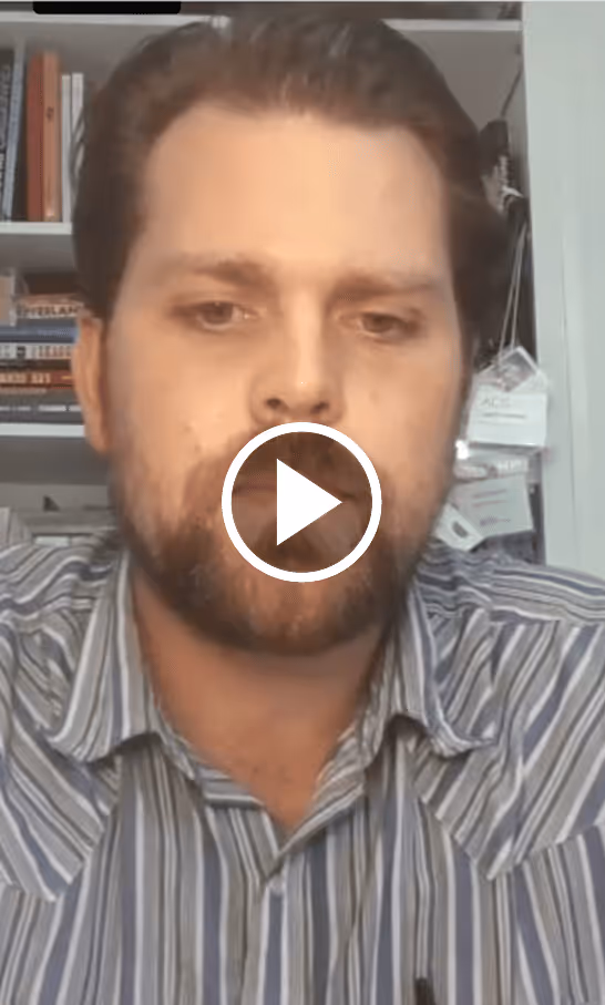 Bearded man wearing a striped shirt seated in front of bookshelves and hanging cards, with a video play button overlay.