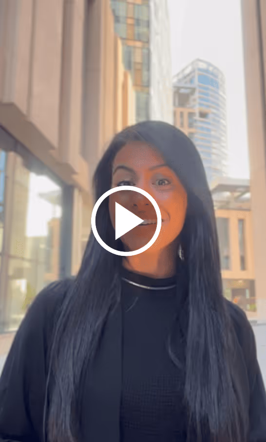 Woman with long black hair smiling outside in an urban area with tall buildings in the background.