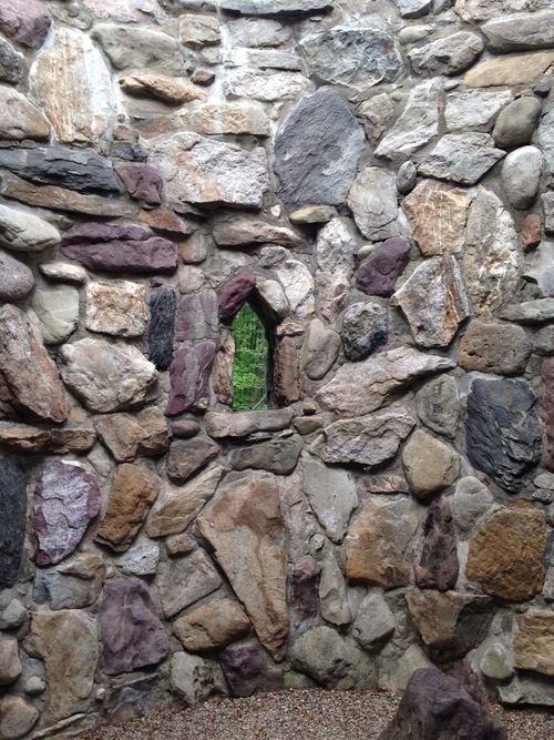 Old stone wall with a small window-like hole through which can see green trees. Rather fae-like