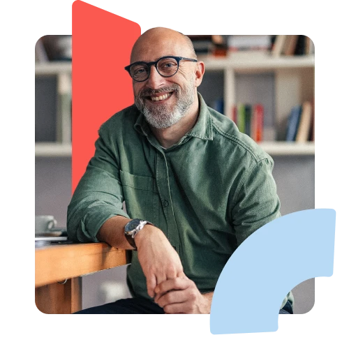 A man wearing glasses is leaning on table cheering at camera. Red shape in the background, blue shape in foreground.