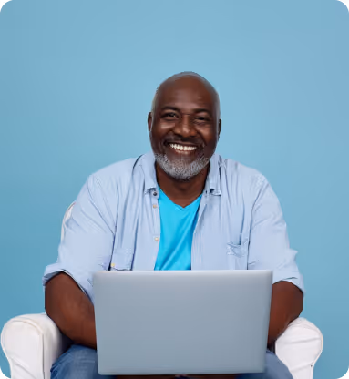 Smiling man with gray beard working on a laptop seated in a white chair against a blue background.