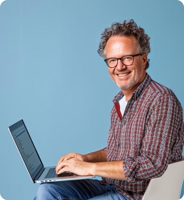 Smiling middle-aged man with curly hair and glasses typing on a laptop against a blue background.