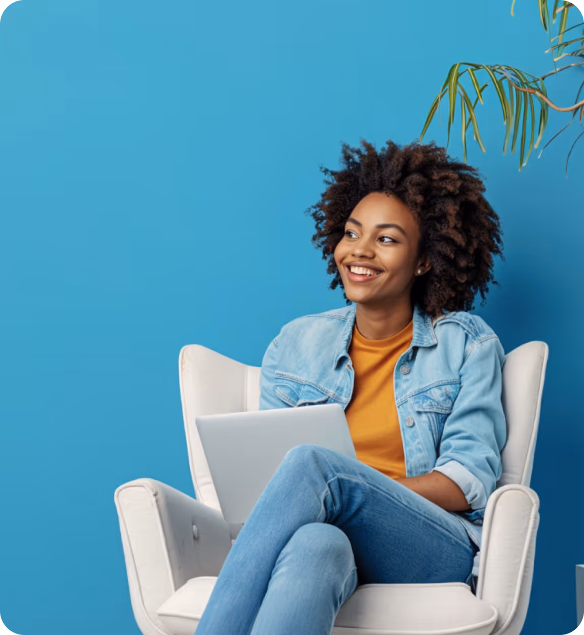 Smiling woman with curly hair sitting in a white armchair holding a laptop against a blue wall.