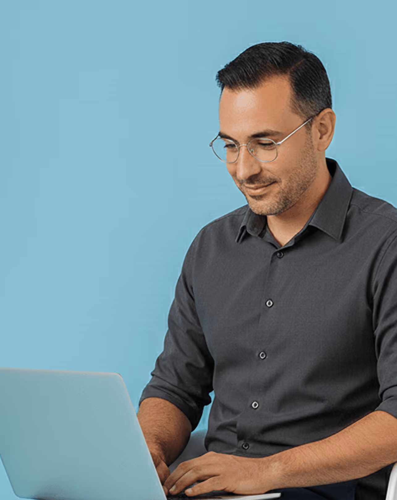 Smiling man with glasses typing on a laptop against a light blue background.