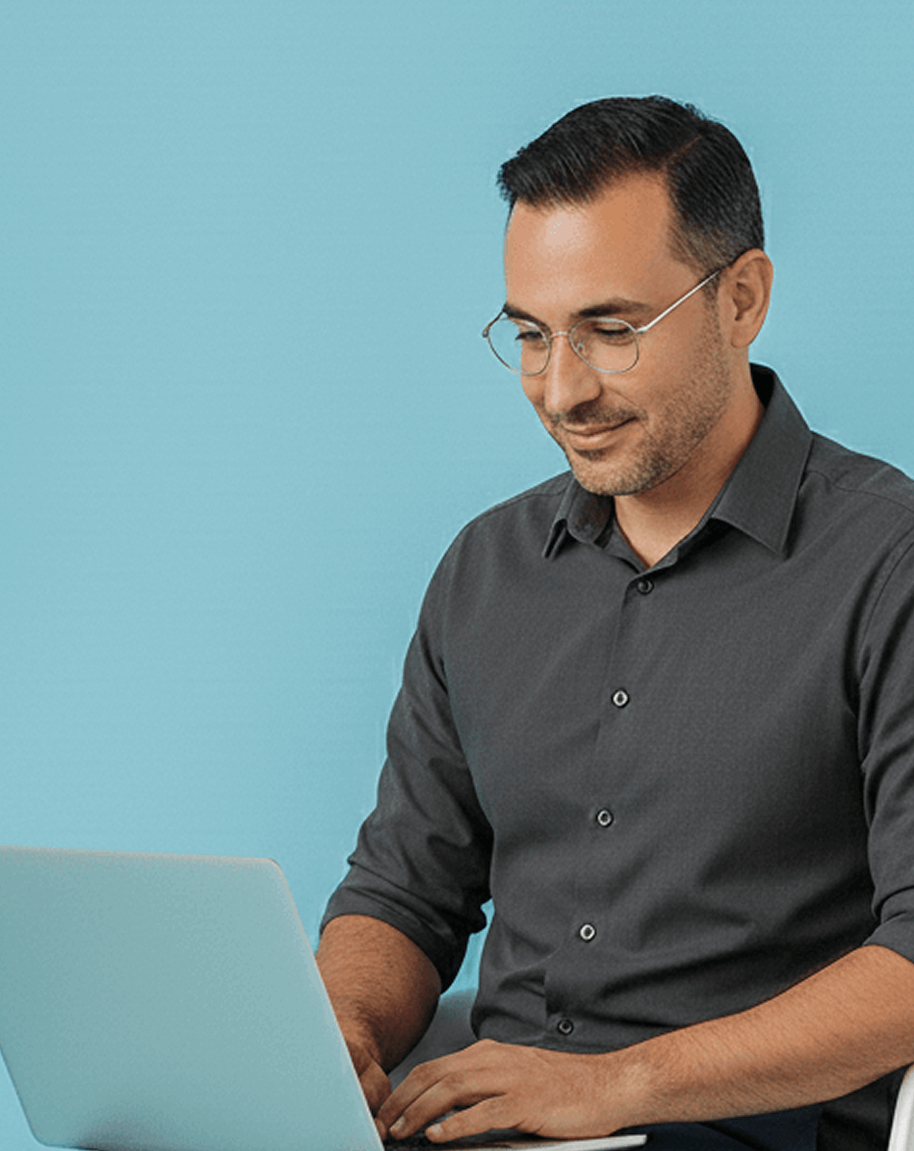 Smiling man with glasses typing on a laptop against a light blue background.