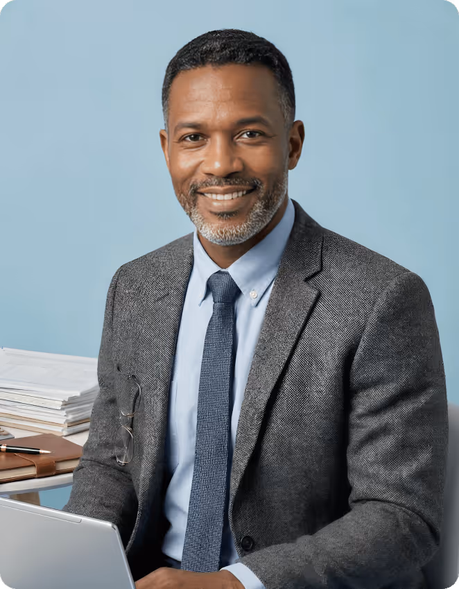 Smiling man in a grey suit jacket and blue tie sitting with a laptop and stack of papers against a light blue background.