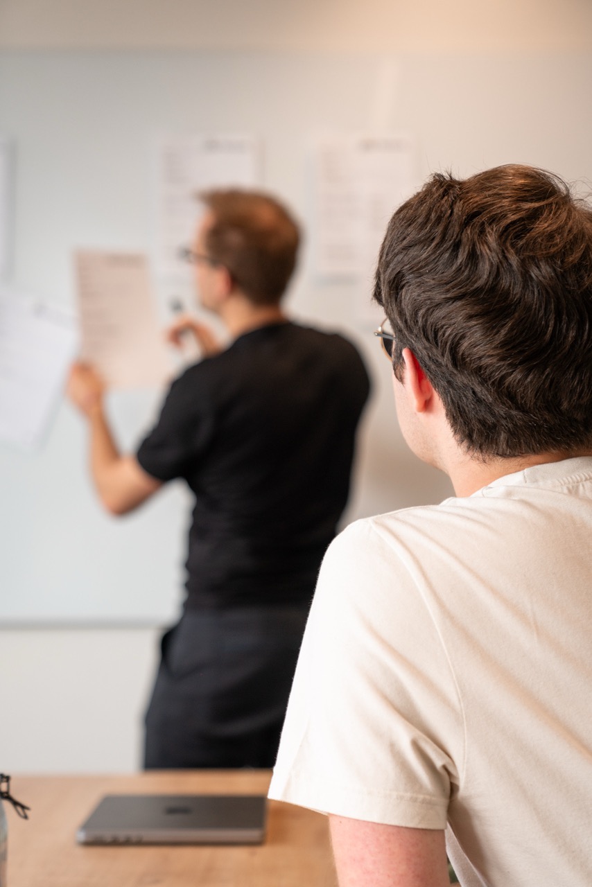 2 people in meeting, 1 at table, 1 at a whiteboard