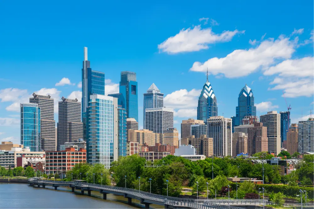 Philadelphia skyline featuring modern skyscrapers, including the Comcast Technology Center and Liberty Place towers, under a bright blue sky with scattered clouds. A river and bridge with green trees in the foreground complete the cityscape.