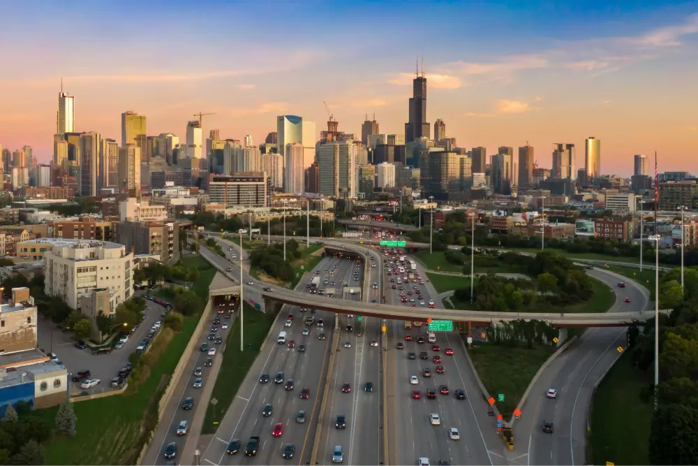 Chicago skyline at sunset with the Willis Tower prominently visible, surrounded by numerous skyscrapers. A busy highway with multiple lanes of traffic and overpasses leads into the city, bordered by green spaces and urban buildings.
