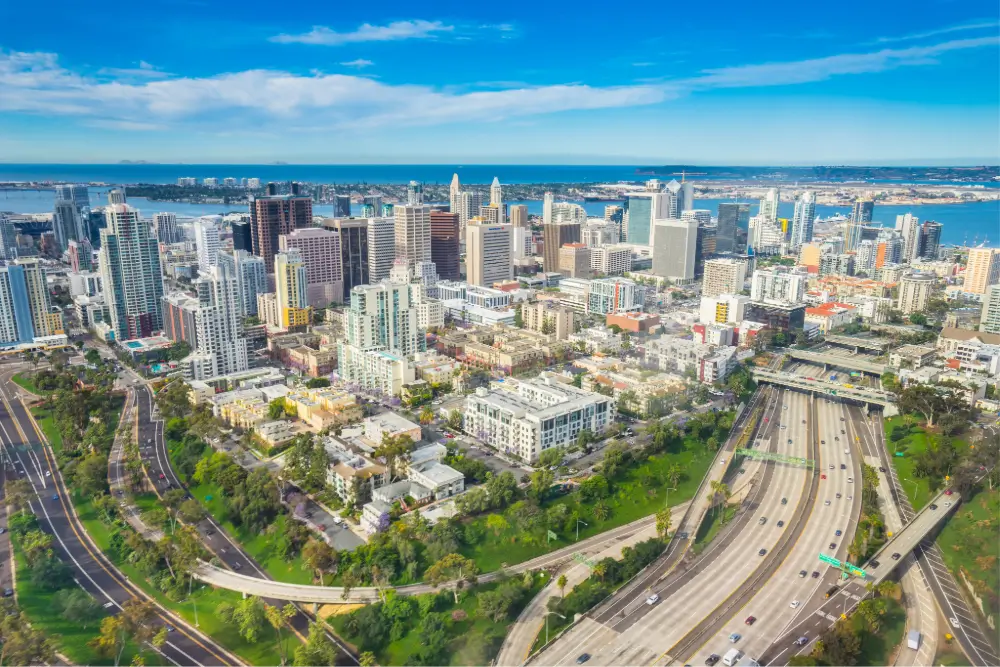 Aerial view of downtown San Diego, California, featuring high-rise buildings, curved highways with light traffic, and green parks. The city is bordered by the blue waters of the bay under a bright, clear sky.
