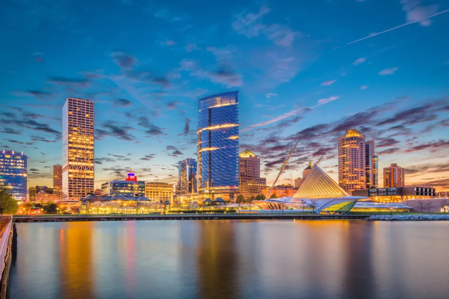 Milwaukee city skyline at dusk with illuminated modern buildings reflecting on calm water.