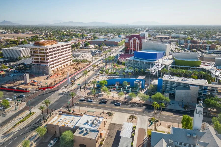 Aerial view of a cityscape in Mesa, Arizona, featuring a mix of commercial buildings, palm trees, roads, and distant mountains under a clear sky.