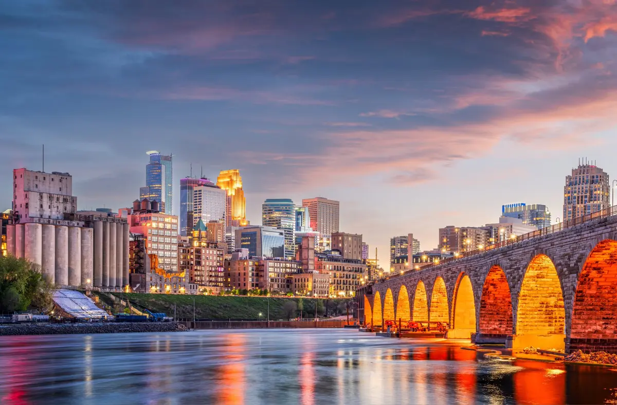 Minneapolis city skyline at sunset with illuminated stone arched bridge over river.