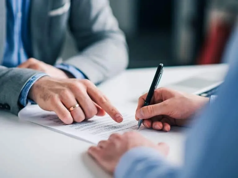 Close-up of a person holding a pen to sign a contract while another person in a suit points to the signature line.