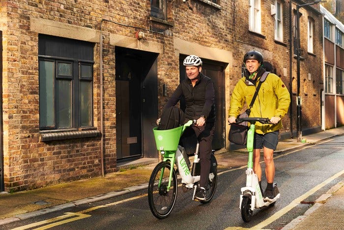 Two men are travelling along street - one is on a white and green branded Lime bicycle and the other on a Lime e-scooter