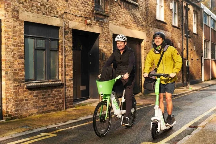 Two men are travelling along street - one is on a white and green branded Lime bicycle and the other on a Lime e-scooter