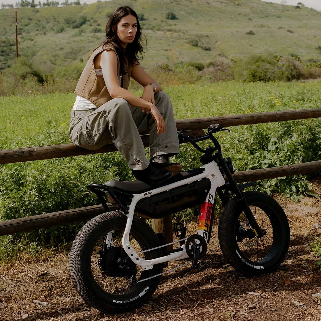 A woman in light colored pants and a light top underneath a tan vest sits on a wooden fence running along a green field. In front of her, a SUPER73-M1D SE is leaning against the fence and the woman is resting her feet atop it.