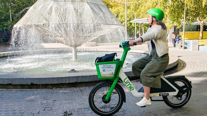 A person rides a green Lime bike near a fountain in an urban space.