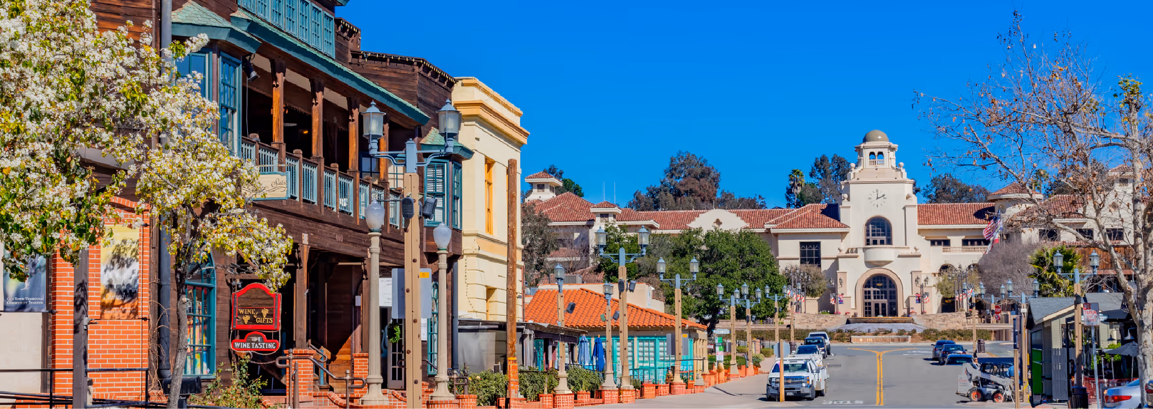 Street view of a charming downtown area with historic buildings, colorful storefronts, blooming trees, and a clock tower under a clear blue sky.