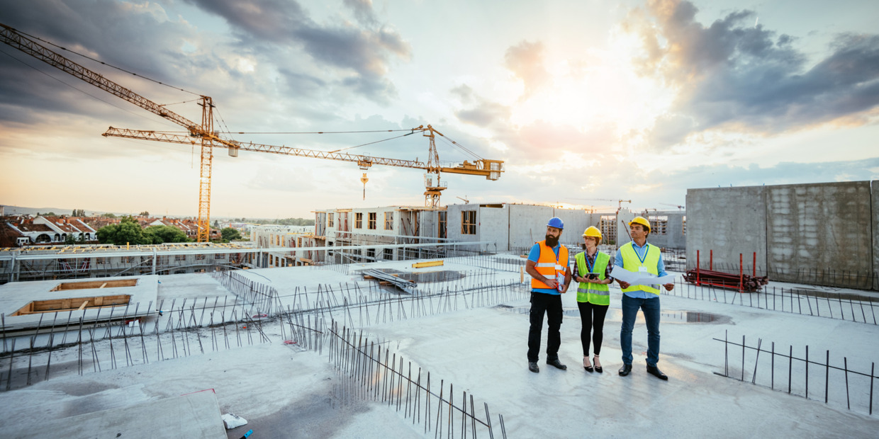 Three construction workers in hard hats and vests review plans on a partially built concrete site with cranes in the background.