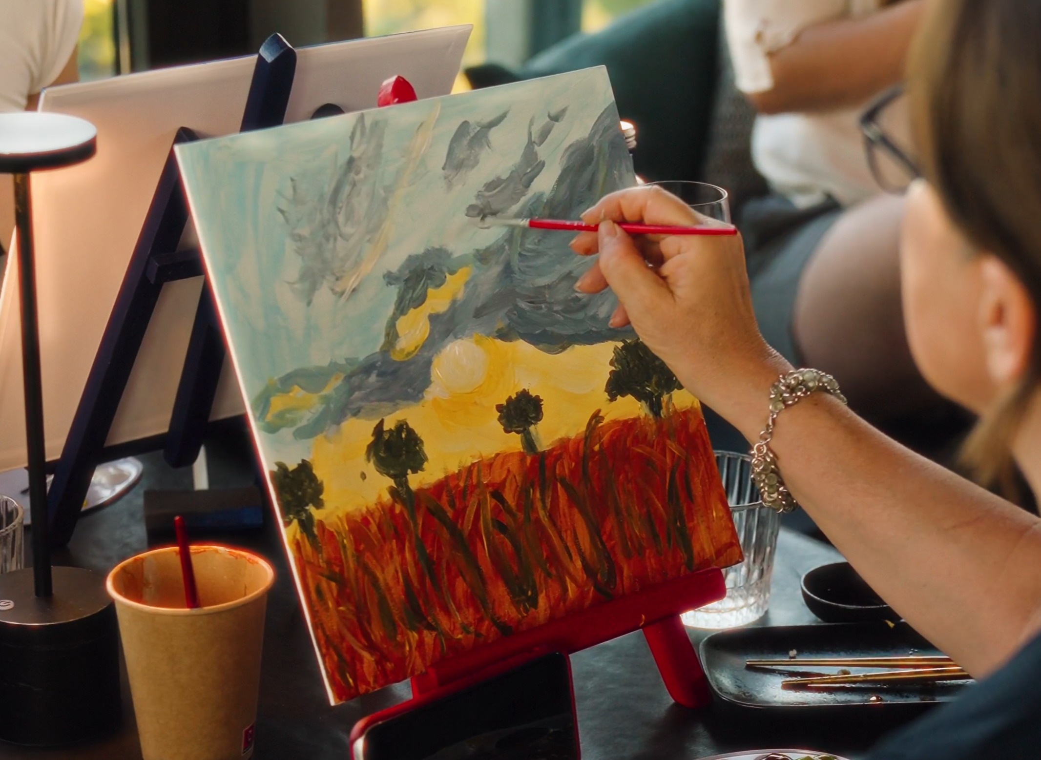 Person painting a landscape with dark clouds, yellow sky, and red grass on a canvas set on a red easel.