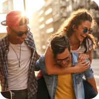 Three people smiling and wearing sunglasses walking in a city. One man is carrying a woman on his back.