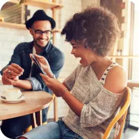 Two people sitting at a cafe laughing while looking at a phone. 
