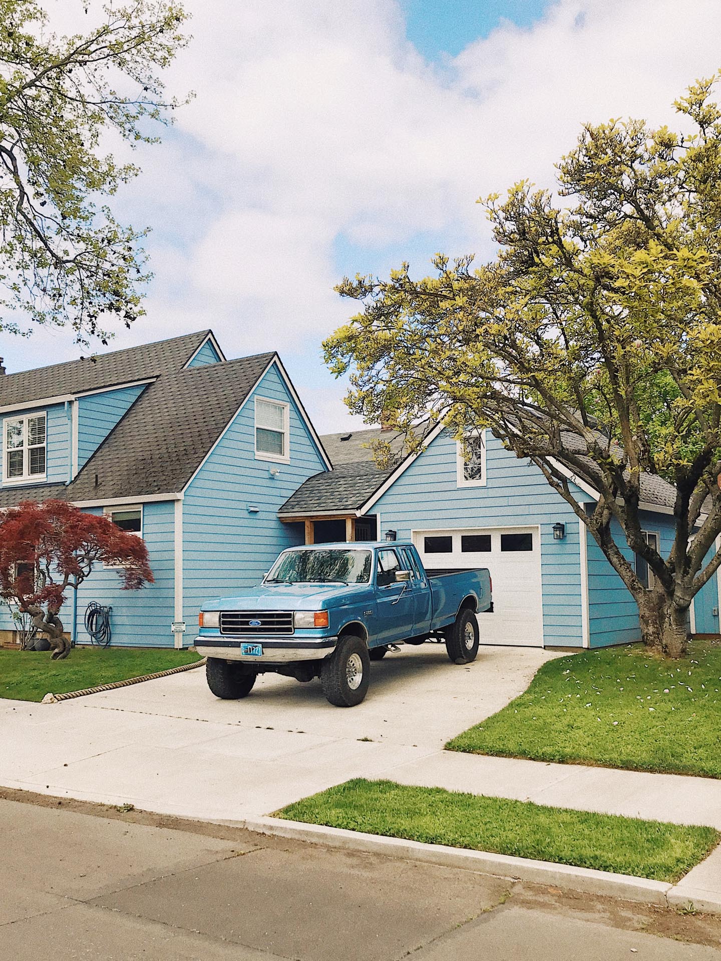 A 90s era periwinkle blue ford truck in the driveway of a light blue house