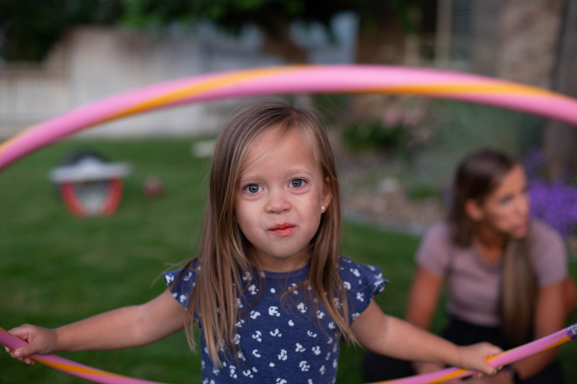 A little girl holds a hula hoop in the front yard with her mom and family behind her
