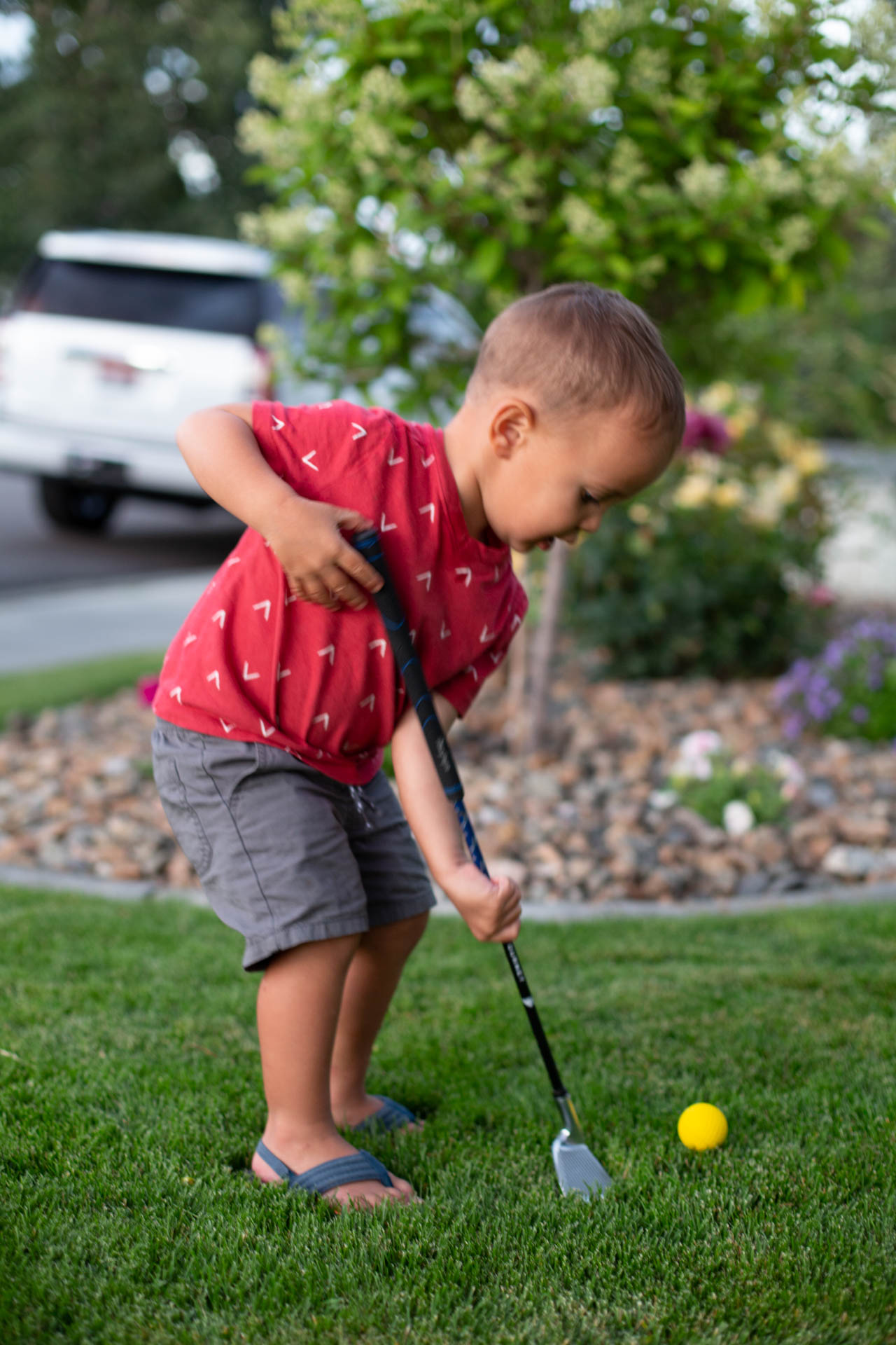 A young boy plays with a golf club and golf ball near a large stone-lined planter in the front yard