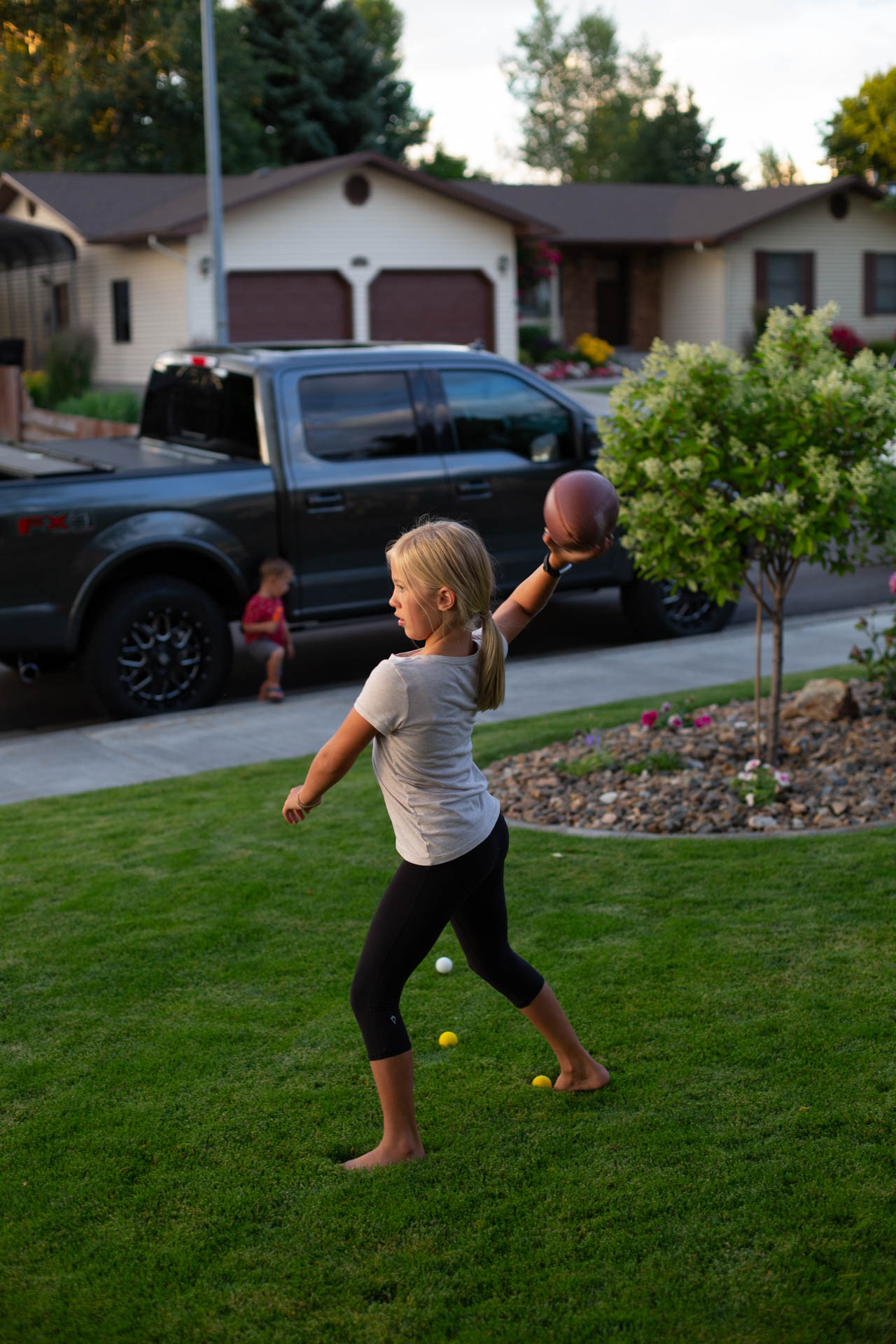 A young blonde girl throws a football in the front yard, with a truck parked on the street in the background