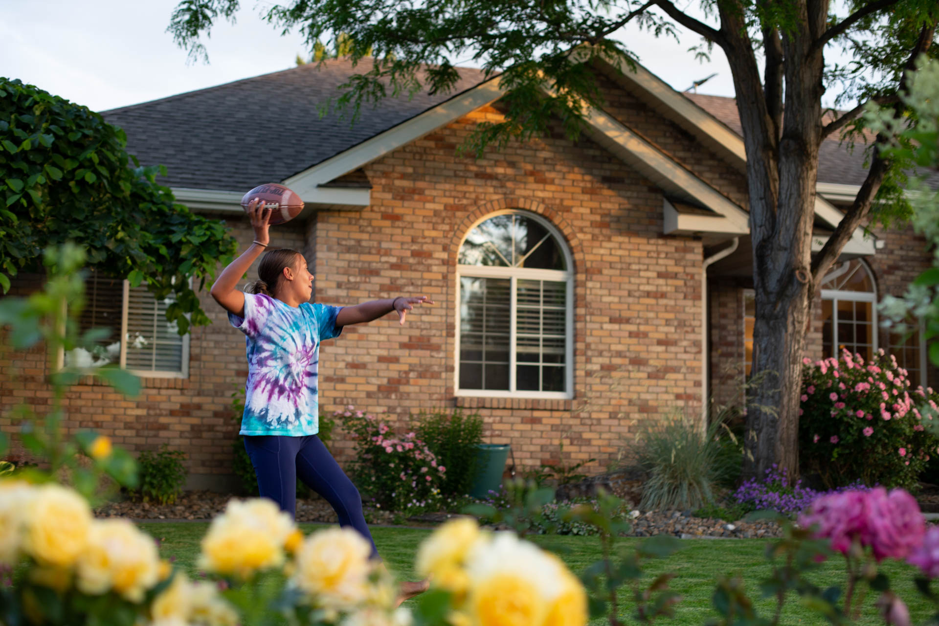 Young brunette girl throws a football in the front yard. Taken from the perspective of the sidewalk, the house is behind her and some yellow and pink flowers are in the foreground