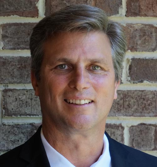 A middle aged caucasian man with hair that is graying on the sides, wearing a white collared shirt with a dark blue blazer